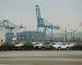A lineup of parked cars at a bustling industrial shipping port with cranes and containers.