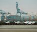 A lineup of parked cars at a bustling industrial shipping port with cranes and containers.