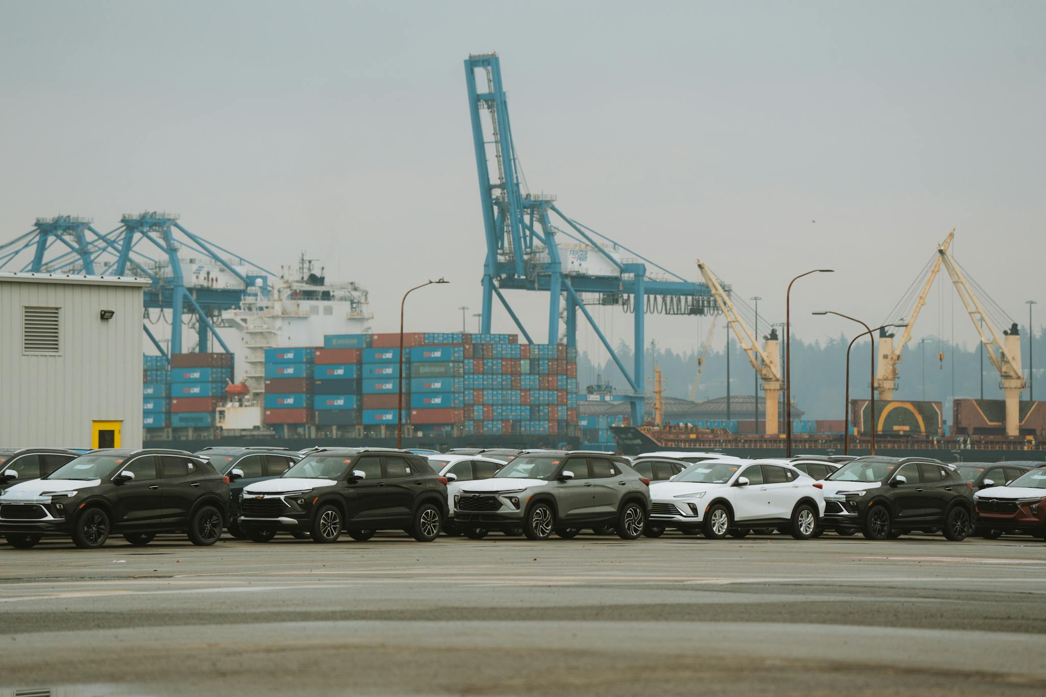 A lineup of parked cars at a bustling industrial shipping port with cranes and containers.